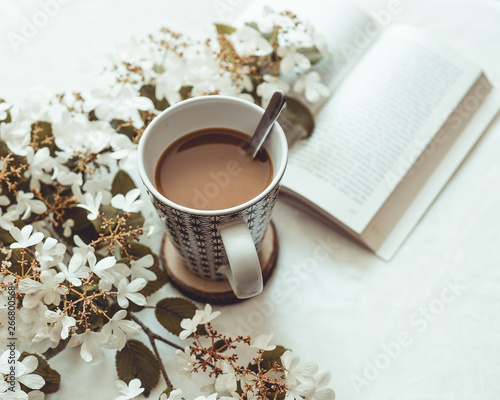 Cup of coffee with milk, with some white flowers in the blur, and an opened book