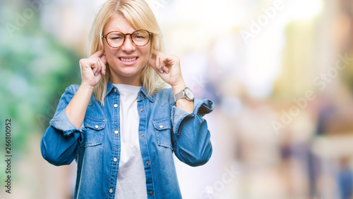 Fototapeta Naklejka Na Ścianę i Meble -  Young beautiful blonde woman wearing glasses over isolated background covering ears with fingers with annoyed expression for the noise of loud music. Deaf concept.