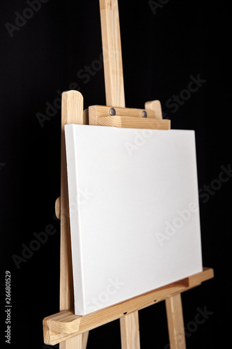 White blank cotton canvas stands on a wooden artistic easel on black curtain background. Horizontal rectangular mockup canvas wrapped on stretcher bar