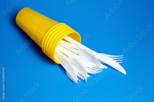 Fototapeta Naklejka Na Ścianę i Meble -  Disposable plastic tableware on blue background. Cutlery, white forks and knives inside yellow cups