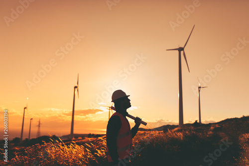 Giovane Ingegnere Aerospaziale con caschetto bianco e gilet arancione, sta controllando il progetto sul foglio di una turbina di un impianto eolico in montagna. Concetto ingegneria ed energia pulita photo