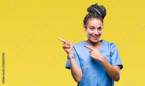 Young braided hair african american girl professional nurse over isolated background smiling and looking at the camera pointing with two hands and fingers to the side.