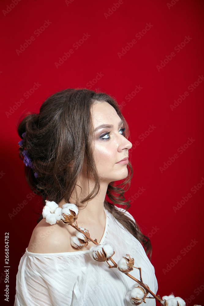 Portrait of young woman with cotton plant with white fluffy flowers on red background. Attractive lady with long wavy brown hair