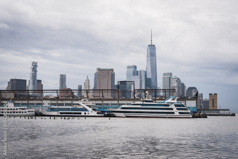 Manhattan skyline with yachts moored at the pier from the greenway of the Hudson River by an overcast blue sky - New York City, NY