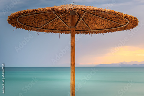 Fototapeta Naklejka Na Ścianę i Meble -  Straw beach parasol, sunset sky and silky, turquoise sea water on Paradisos beach in Neos Marmaras, Greece