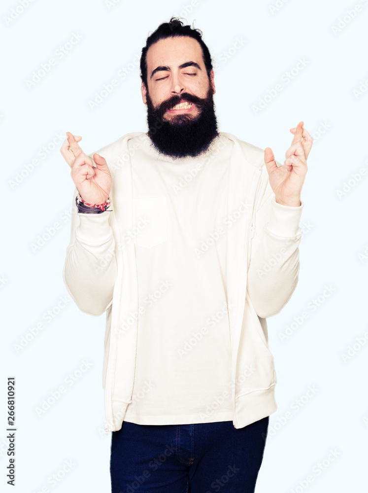 Fototapeta premium Young man with long hair and beard wearing sporty sweatshirt smiling crossing fingers with hope and eyes closed. Luck and superstitious concept.