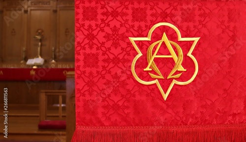 Close-up of red lectern parament with  first and last letters of Greek alphabet in gold. Concept that God is the beginning and end of all things. Blurred altar, cross, Bible and candles in  background photo