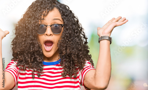 Young beautiful woman with curly hair wearing sunglasses celebrating crazy and amazed for success with arms raised and open eyes screaming excited. Winner concept