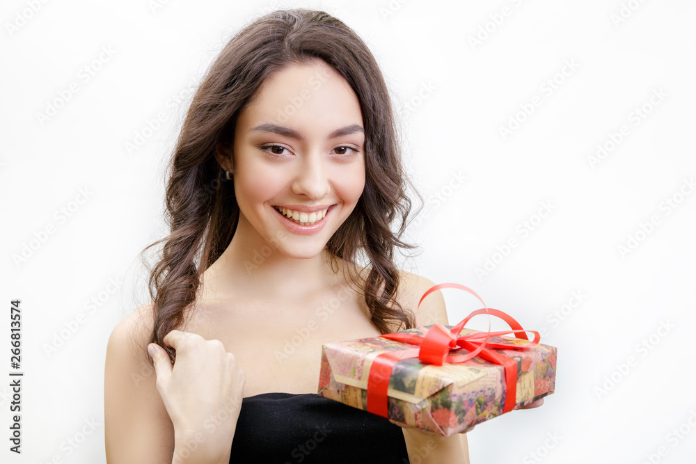 Beautiful young cheerful woman holds gift with a red bow. She looks at the camera and smiling. Girl has brown medium hairstyle with curls. Isolated on white background. Beautiful young cheerful woman holds gift with a red bow. She looks at the camera and smiling. Girl has brown medium hairstyle with curls. Isolated on white background.