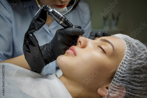 Permanent make up eyeliner procedure, applying on young girl. Close-up. Young beautiful woman making permanent makeup in cosmetology salon. Young girl applying permanent eyeliner in beauty studio