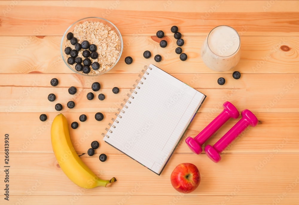 Glass of yogurt, banana, blueberries, apple, oat flake, notebook and dumbbells on wooden background. Glass of yogurt, banana, blueberries, apple, oat flake, notebook and dumbbells on wooden background.