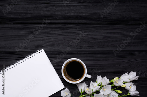 White flowers bells, coffee and empty notebook for your text on a black wooden background.