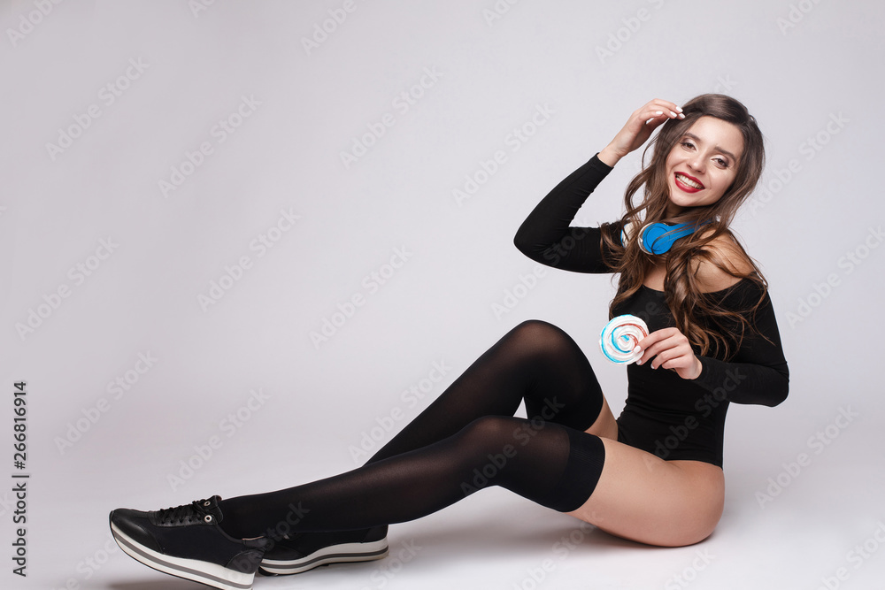 View from side of young girl wearing black sneakers, stockings and body and  listening music in headphones. Happy female keeping candy, looking at  camera, posing and smiling on isolated background. Photos