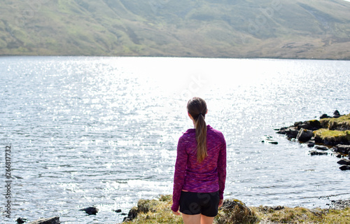 Brown haired girl in purple jumper looking out of sunny tarn/ mountain lake in Grisedale pike, Lake District, England, UK photo