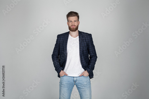 A beautiful sexy bearded man in a jacket holds his hands on his jeans and looks at the camera. he stands in front of the white background.
