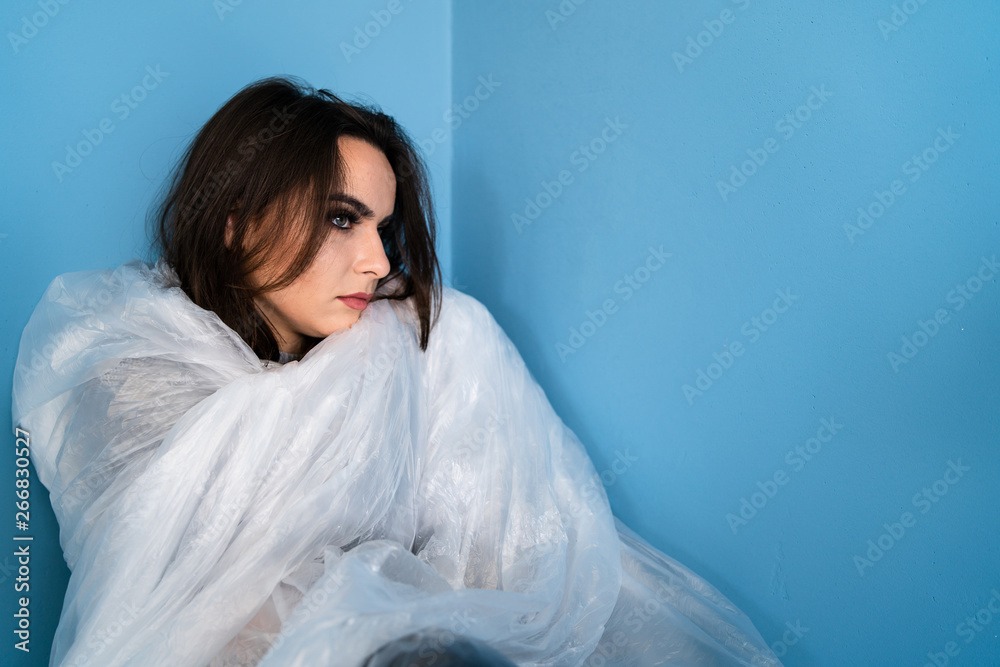 Young scared woman sad covered with plastic nylon cellophane sheet sitting helpless in a corner of a blue room trapped human trafficking anti violence pollution save the earth concept Young scared woman sad covered with plastic nylon cellophane sheet sitting helpless in a corner of a blue room trapped human trafficking anti violence pollution save the earth concept