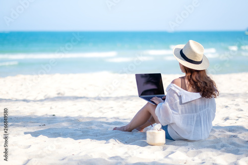 A woman using and typing on laptop computer keyboard while sitting on a beautiful beach