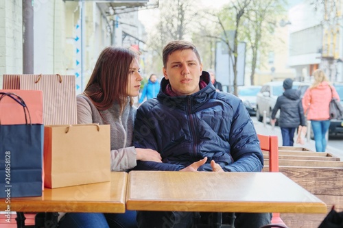Young tired couple with shopping bags in street cafe, waiting for cup of coffee and tea