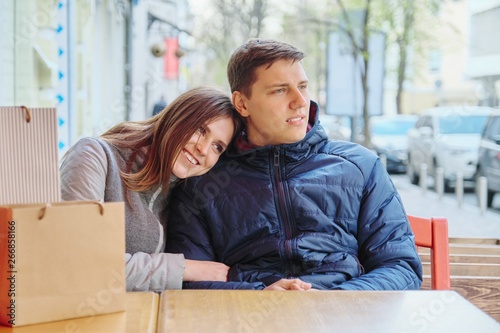 Young smiling couple with shopping bags in street cafe, waiting for cup of coffee and tea