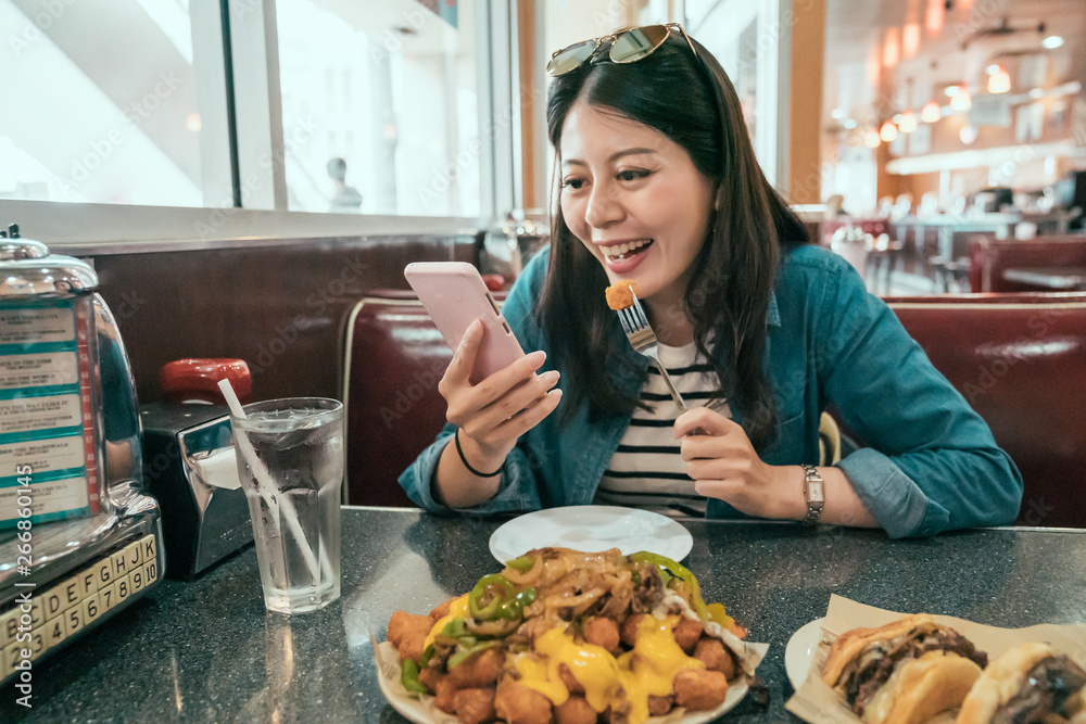 vintage style young asian girl traveler sitting in cafe and drinking soda cold in glasses with ice while using mobile phone. happy laughing local woman resting in diner in afternoon eating fast food