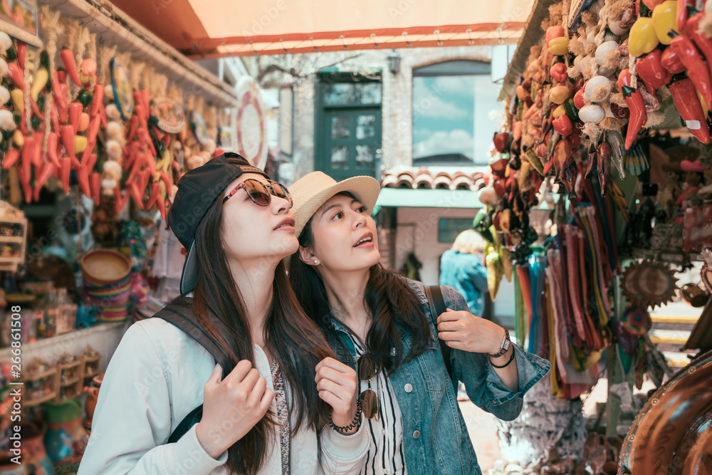 two asian young women on summer vacation walking in outdoor mexican  market. smiling friends ladies shopping in local vendor selling decor chilli for home in olvera street. girls look up together.