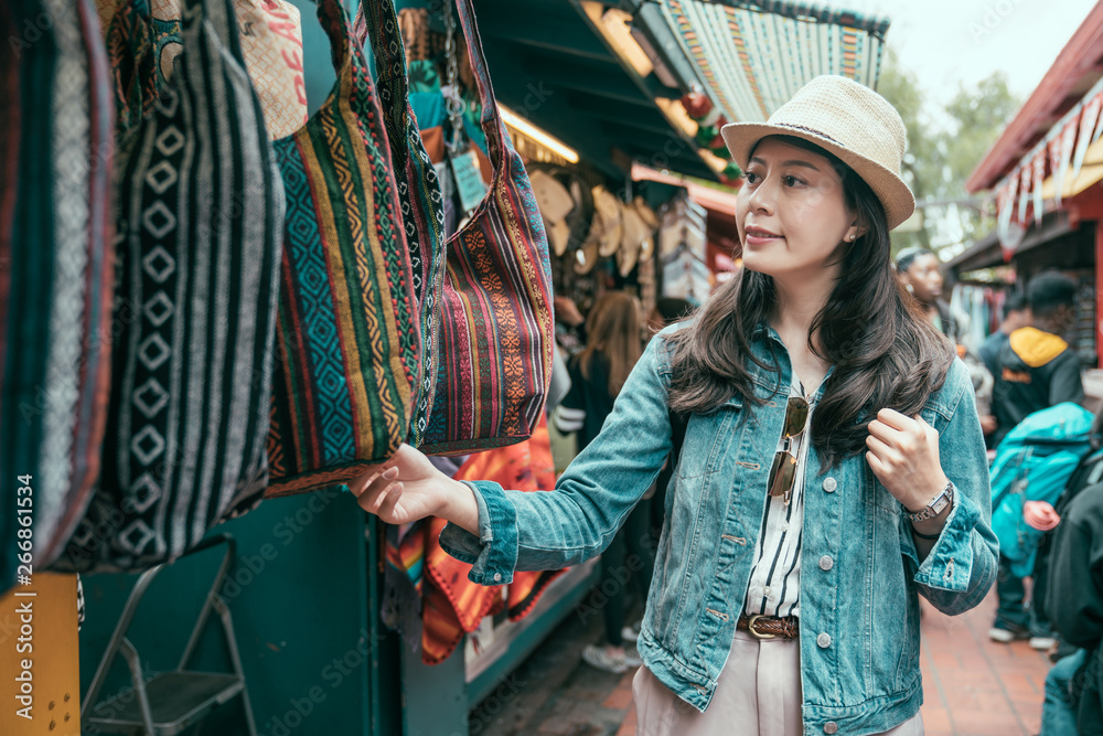 Summer holidays shopping concept. Young asian woman tourist buying souvenirs in gift olvera street stall outdoor picking choosing bags at vendor. girl traveler shopping in mexico market on sunny day