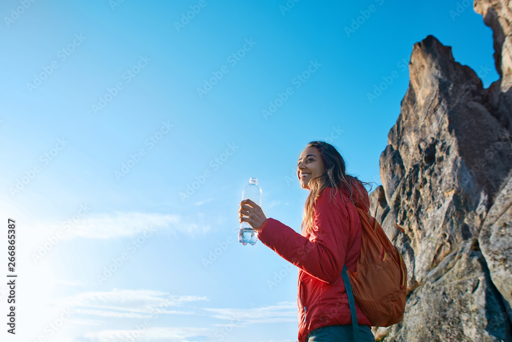 woman hiker with backpack, wearing in red jacket, standing on edge of cliff against a blue sky background. Young female holds a bottle of water in hand and enjoying.