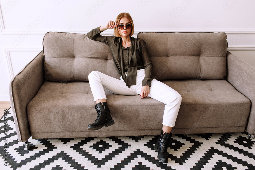 Obraz premium Beautiful sweet girl lying on the sofa. Golden stylish model with short red hair, plump lips, sunglasses, casual clothing, costume, outfit, overalls. Model test, advertising glasses, clothes