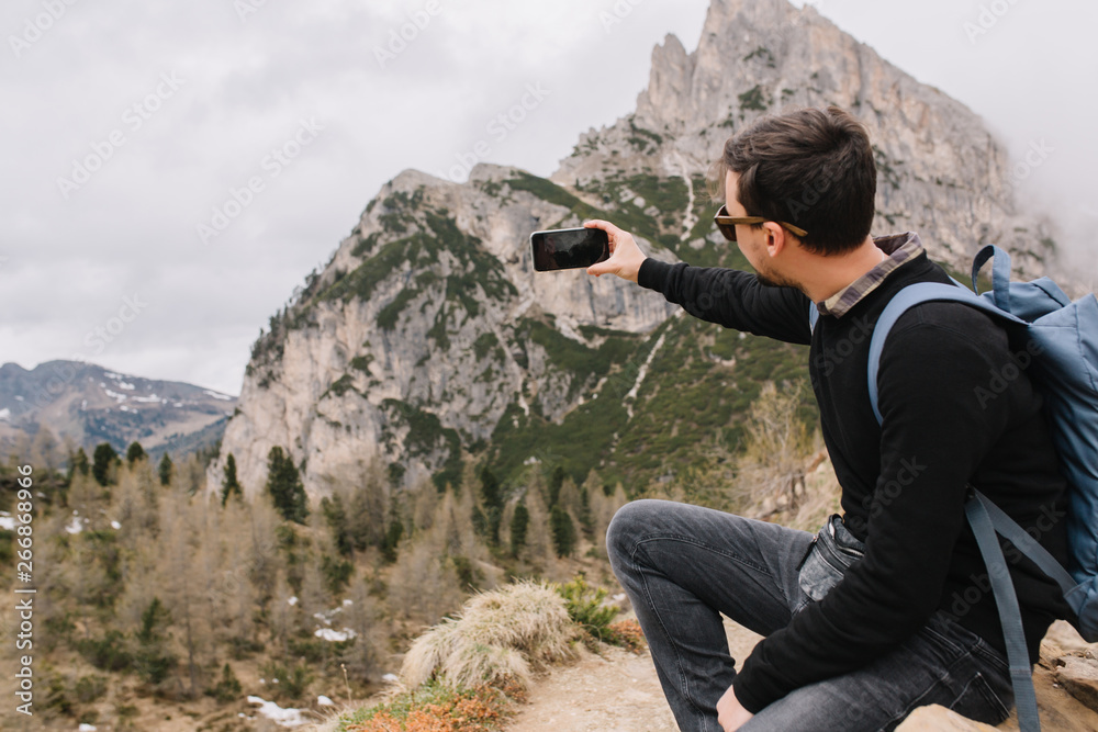 Young man with dark short hair sitting on the stone under overcast sky and looking at rocky landscape. Boy wearing black shirt resting after climbing and taking picture of italian mountains.