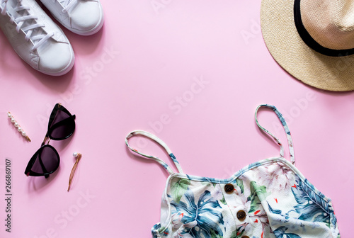 Top view of a woman's summer clothes for city street look. Exotic printed summer dress with straw hat, white sneakers and trendy sunlasses on pink background.
