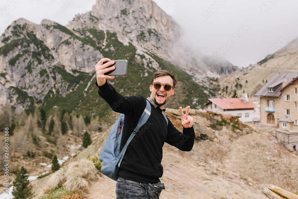 Obraz premium Excited young man with short haircut posing in mountains with cozy house on background. Outdoor portrait of traveler with backpack in trendy sunglasses making selfie during trip around Italy.