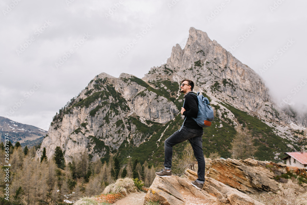 Handsome brunette man stands on the rock admiringly looking at amazing nature views. Outdoor full-length portrait of active tourist spending time in mountains and climbing to the top.