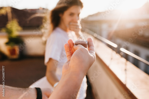 Man holding wedding ring in front of astonished happy girl covering mouth with hand. Romantic photo of charming woman standing on roof early in evening on date with boyfriend in anniversary. photo