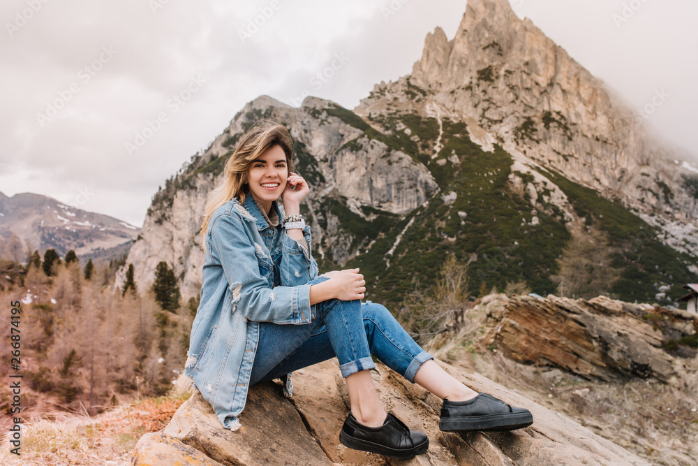 Inspired smiling girl wearing black leather shoes chilling on stone after long trekking and posing with pleasure. Portrait of adorable female traveler addicted to active tourism in mountains.