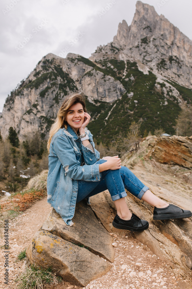 Naklejka premium Stunning blonde lady posing on stone spending time in italian Alps in cloudy spring morning. Outdoor portrait of graceful cute girl travelling in mountains in vacation and posing on amazing landscape.