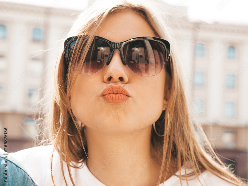 Portrait of beautiful smiling blond model dressed in summer hipster  clothes. Trendy girl posing in the street background. Funny and positive woman in sunglasses. Making duck face