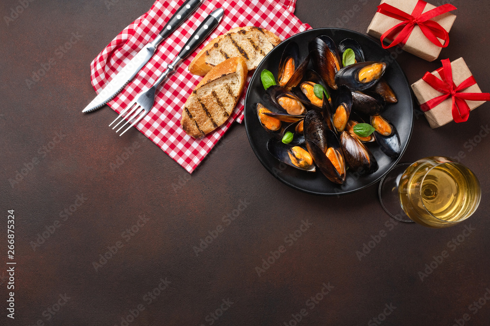 Seafood mussels and basil leaves in a black plate with wineglass, fork, knife and gift boxes on towel and rusty background