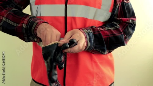 Male worker dressed in a reflective vest put on blue waterproof protective gloves, show thumbs up, sign OK. Hands closeup isolated on light green background.