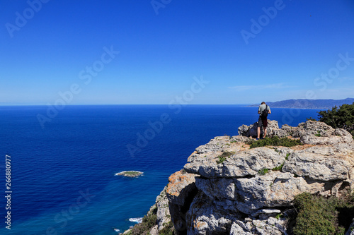 Back view of freedom man stand on rock cliff looking at blue sea and Cypriot nature. Contrast between midget and infinitely water land. Rock hill in Akamas peninsula national park with athlete