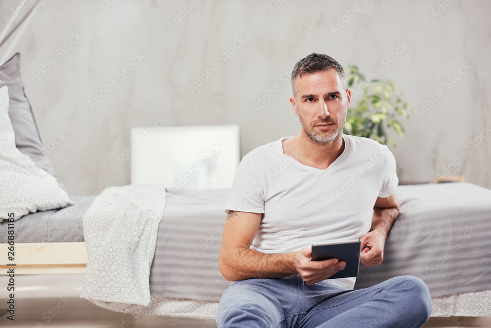 Handsome Caucasian middle-aged man sitting on the floor in bedroom and leaning on bed. In holding hands tablet.
