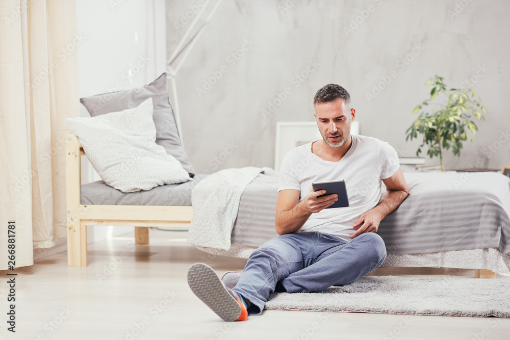 Handsome Caucasian middle-aged man sitting on the floor in bedroom and leaning on bed. In holding hands tablet.