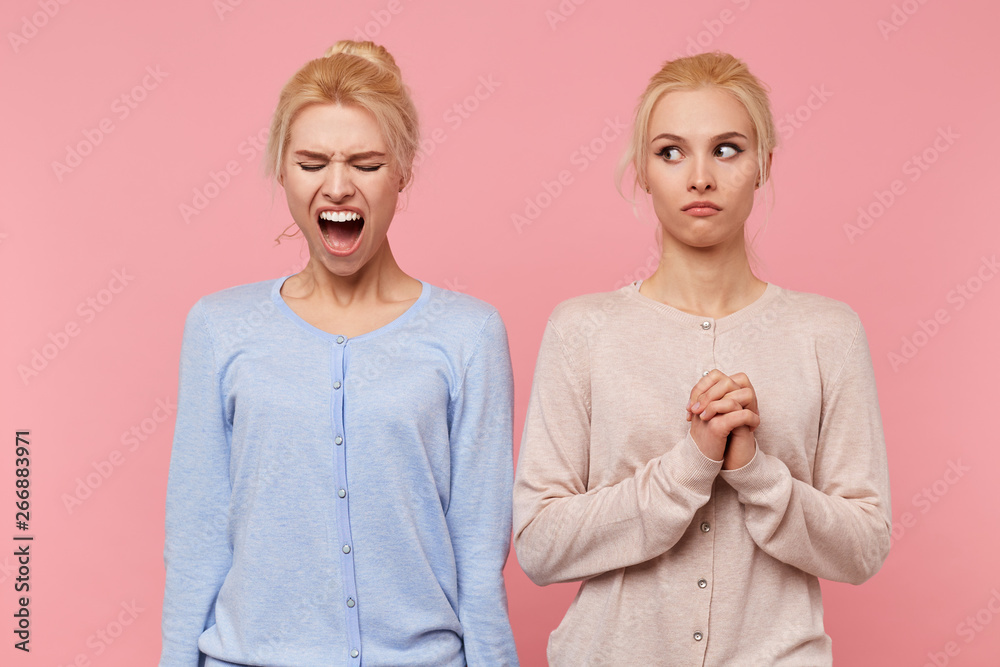 Young cute blonde woman frightened looks at her screaming sister isolated over pink background, people and emotions concept. Young cute blonde woman frightened looks at her screaming sister isolated over pink background, people and emotions concept.