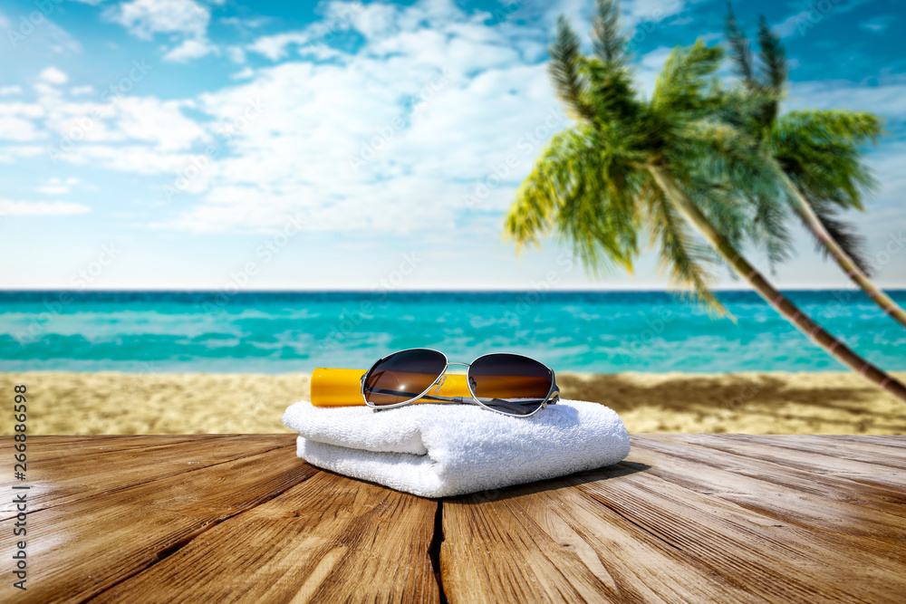 Wooden table background of free space for your decoration. White towel background with sunglasses and beach landcsape with palms and ocean. Blue sky with sun light. Summer time on beach. 