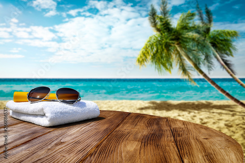 Wooden table background of free space for your decoration. White towel background with sunglasses and beach landcsape with palms and ocean. Blue sky with sun light. Summer time on beach.  #266886537