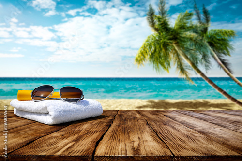 Fototapeta Naklejka Na Ścianę i Meble -  Wooden table background of free space for your decoration. White towel background with sunglasses and beach landcsape with palms and ocean. Blue sky with sun light. Summer time on beach. 