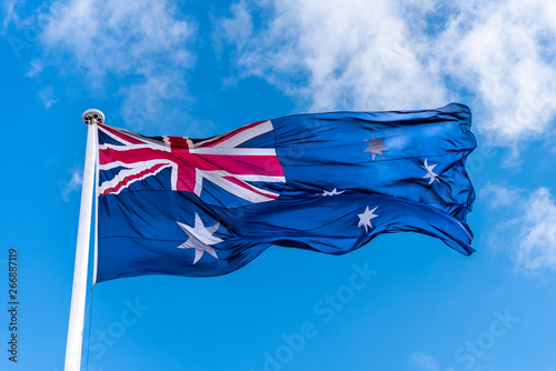 The Australian flag attached to a white flag pole, flying in the wind against a light cloud blue sky