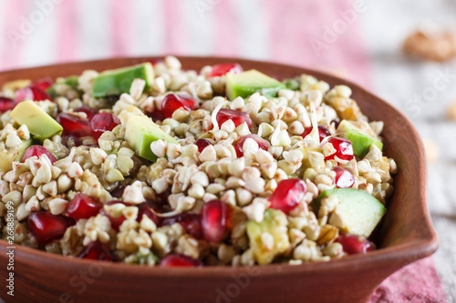 Salad of germinated buckwheat, avocado, walnut and pomegranate seeds in clay plate on white wooden background.