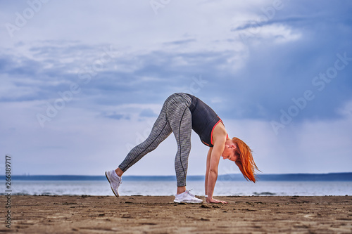 A middle-aged woman with red hair does gymnastics on the sandy shore of a large river. Cloudy spring morning.