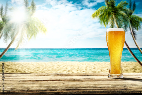 Summer background of wooden desk and cold beer. Free space for your decoration. Beach landscape with palms and ocean. Summer sunny day with sun light. Beach party and summer time. 
