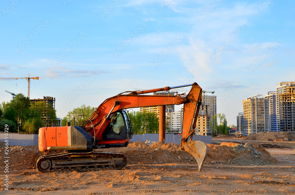Heavy tracked excavator at a construction site on a background of a residential building and construction cranes on a sunny day against the backdrop of a sunset and blue sky with clouds